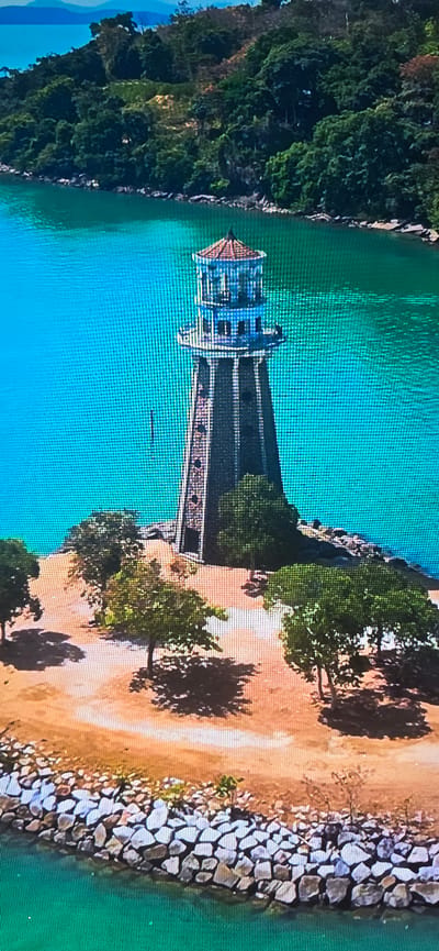 The image depicts a lighthouse situated on a small, sandy island. The lighthouse is a tall, cylindrical structure with a dark, possibly stone or brick, base and a lighter-colored upper section. It has several levels, with windows or openings at different heights, and a lantern room at the very top, topped by a conical roof. The island is sparsely populated with trees, some of which cast shadows on the sandy ground. The foreground is a rocky shoreline, where light-colored stones are stacked to form a barrier. The island is surrounded by bright, turquoise water, and in the background, lush green hills or mountains covered in dense vegetation can be seen. The sky above is a muted blue. The image is captured from a high vantage point, suggesting an aerial perspective. There are no people visible in the image. The scene is serene and natural, with the lighthouse standing as a solitary structure against the backdrop of the sea and the verdant landscape. It is not possible to determine the location in the city of Lippstadt, Germany, as the image does not contain any identifiable landmarks or contextual clues to suggest this specific location.