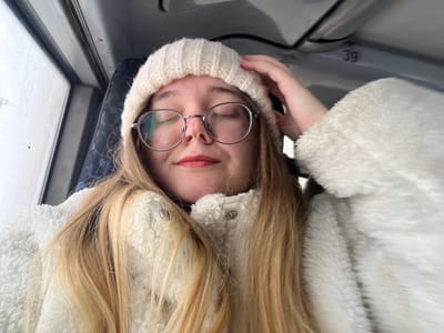 A young adult female, with long blonde hair, fair skin, and round wire-rimmed eyeglasses, is depicted inside what appears to be a public transport vehicle in the city of Липицы, Russia. She is wearing a cream-colored knit beanie and a white, textured, possibly sherpa-style jacket. Her eyes are closed, and her lips are slightly parted in a subtle smile, adorned with light pink lipstick. Her right hand is partially visible, touching the top of her beanie on the right side of her head. The background consists of a dark blue patterned seat and a window to her left, which is obscured by condensation or frost, showing an indistinct bright, possibly wintery, outdoor environment. Above her head, a section of the vehicle's gray interior ceiling or overhead compartment is visible, bearing the alphanumeric marking "3g".