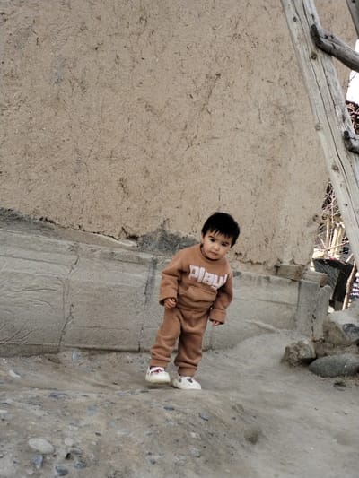 A young child stands on an uneven dirt and gravel surface in Limbur, Uzbekistan. The child, with dark hair and eyes, directly faces the viewer. The individual wears a light brown two-piece tracksuit with "PLAY" in white capital letters on the sweatshirt's front pouch pocket, and white sneakers featuring a small red accent on the left heel. Behind the child is a weathered, light brown wall, possibly mud or adobe, exhibiting a textured surface and a prominent concrete or stone base. A rustic wooden structure, resembling a ladder, leans against the wall on the right side of the frame, with bare tree branches visible beyond. The scene is illuminated by diffuse natural light.