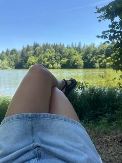 A person's legs, wearing a light blue denim skirt and black sandals, are crossed as they relax on the grassy bank of a lake. Lush green trees form a dense forest lining the far side of the lake, under a clear blue sky. The water appears a murky green, reflecting the surrounding foliage. The scene suggests a peaceful outdoor setting.