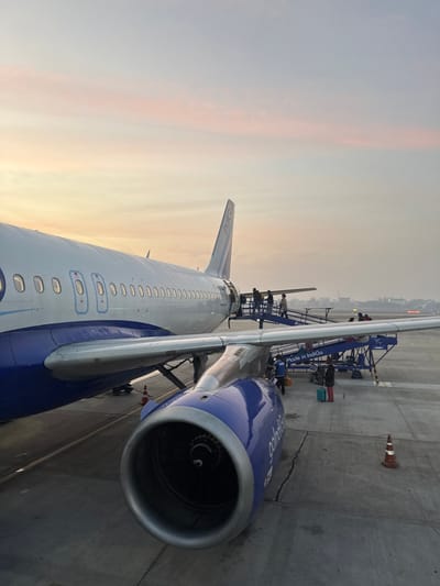 A vertically-oriented shot depicts the wing and engine of a blue and white Indigo airplane. Visible are several maintenance workers on scaffolding near the wing's root, a safety cone sits on the concrete tarmac. The fuselage features a row of oval windows. The sky has shades of yellow and orange.