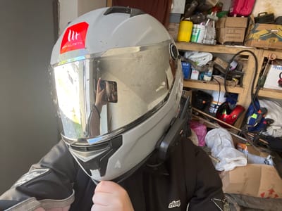 Here's a detailed description of the image:
**Scene:** The image appears to be taken in a cluttered garage or storage area. Various objects are visible in the background, including boxes, containers, bottles, tools, and what looks like another helmet.
**Subject:** The primary subject is a gray and silver motorcycle helmet held up by a person wearing a black and gray jacket. The helmet has a reflective visor and a red sticker with the brand "MT Helmets". The visor is dirty with smudges and bugs.
**People:** A hand and part of a jacket are visible, suggesting a person holding the helmet. The reflection in the visor of the helmet shows the image being taken on a cell phone.
**Environment:** The background is filled with clutter, indicating a possibly disorganized storage space.
**Location:** The location is not determinable from the image. Cherliany, Ukraine is not determinable from the image.