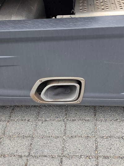 A close-up, eye-level shot shows the exhaust pipe of a truck. The exhaust pipe is rectangular with rounded corners and has a silver, metallic finish. It is embedded in the dark gray side panel of the truck, which has a matte texture and some visible scuff marks. The exhaust pipe is slightly recessed and secured by four screws around its outer rim, which has a slightly discolored, bronzed appearance. The background consists of a textured gray concrete surface with visible pebbles and lines indicating paving stones, consistent with a street or parking lot. The image was taken outdoors. This location is in Zeppelinheim, Germany.