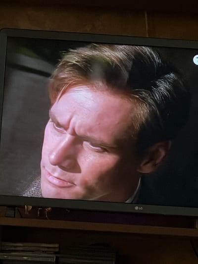 A close-up shot captures the face of a man with light skin and brown hair, looking to his left. His forehead is furrowed, and his lips are slightly parted. He wears a tweed jacket. The background is dark, with a hint of green on the left side. A white circle with a 'T' inside is visible in the upper right corner of the screen. The LG logo is present on the bottom edge of the screen. The image appears to be a still from a television or video recording. There is no indication of the city of Osolynka, Ukraine, in this image.
