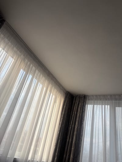 This image depicts an interior view of a room in Tsenovo, Bulgaria, specifically focusing on a corner where two walls meet, both featuring windows covered by curtains. The ceiling, painted a light, neutral color, occupies the upper right portion of the frame. Along the top edge of both walls, curtain tracks are visible, from which curtains are suspended. On the left wall, translucent white sheer curtains (tulle) hang, allowing bright daylight to filter through from outside, creating vertical streaks of light and shadow. Behind these sheer curtains, the outline of windows is discernible. Adjacent to this, in the corner, darker, heavier drapes with a geometric pattern (possibly triangular or abstract shapes) are partially drawn open. Similar sheer white curtains cover the window on the right wall. No people, subjects, or actions are present in the scene, which is illuminated by natural light entering through the windows.