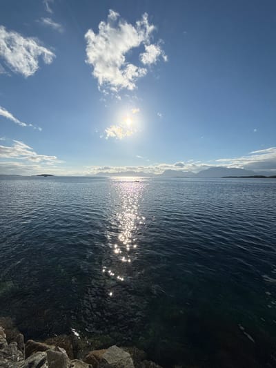 A wide-angle, high-angle shot captures a sunlit body of water with a rocky shoreline in the foreground. The sun is high in the bright blue sky, partially obscured by scattered white clouds, casting a shimmering reflection on the water. In the distance, a chain of mountains forms the horizon, with a small island visible to the left. Two kayakers are present in the water, their figures silhouetted against the sun's glare. The water is clear and dark blue, with visible rocks and seaweed beneath the surface near the shore. The overall scene suggests a calm and bright day, possibly in the vicinity of Harstad, Norway, given the mountainous coastal landscape.