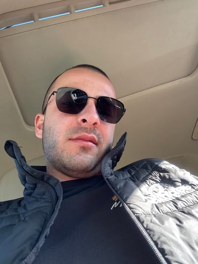 The image shows a man wearing sunglasses and a black puffer jacket, angled upwards towards the car's interior roof. The interior roof appears to be beige in color. No information suggests the location is in Pirdop, Bulgaria.