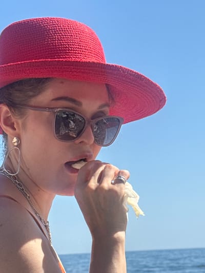 A medium shot captures a woman wearing a red woven hat and sunglasses against a clear blue sky and the ocean. She is in the process of eating something, with her hand holding a piece of food to her mouth. Her left hand, adorned with a ring, is also near her mouth. She wears a silver necklace and a large, abstract silver earring in her left ear. The background shows the ocean and sky, suggesting an outdoor setting, possibly a beach or seaside location. The woman's face is partially obscured by her sunglasses. The overall scene appears casual and summery. There is no indication of the city of Ut'os, Ukraine.