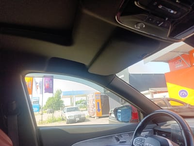 The image is taken from inside a vehicle, looking out through the windshield and side window. It depicts a view of a gas station and surrounding area in Ötegen Batyr, Kazakhstan.

In the foreground, the interior of the car is visible, including the steering wheel and dashboard. The side window looks out onto an exterior scene.

Outside, a white Mercedes-Benz sedan is parked on the left. To its right, there is a yellow kiosk with an orange sign that says "ВХОД ЗДЕСЬ" (Entrance Here) and an arrow pointing down. Behind these, a gas station canopy with the word "AVTOGAZ" in blue lettering is visible. Several flags with "GT OIL SERVICE" logos are planted on the left side of the image. In the background, more vehicles are parked, and trees and buildings can be seen. The sky is bright, suggesting daylight.