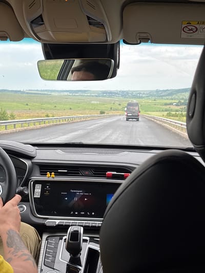 The image is taken from inside a car, looking forward through the windshield. The driver's hands are visible on the steering wheel. The dashboard features a large touchscreen display showing song information. Two small yellow duck figures are perched on top of the air vents. Outside the car, a road stretches into the distance, with a dark-colored van driving ahead. The landscape consists of rolling green hills and fields under a cloudy sky. There is a guardrail along the side of the road. The air vent at the top of the windshield has a sticker indicating "AIRBAG." The rearview mirror reflects the driver's face and hair. This scene is likely taking place on a road near Verkhnyaya Toima, Russia, given the typical landscape of the region and the Russian text visible on the car's infotainment system.