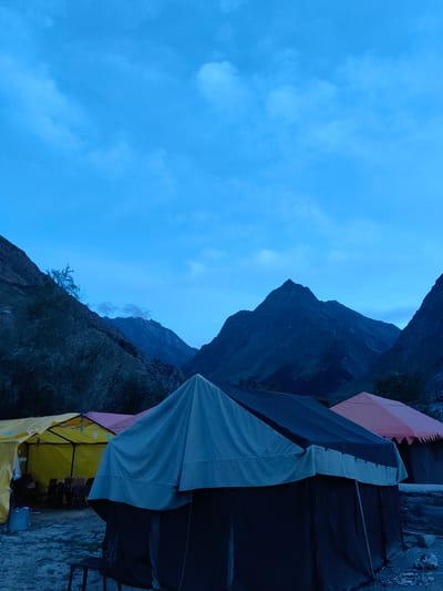 In Gushal, India, at twilight, a campsite is depicted with several tents and mountains in the background. The sky is a deep blue with scattered clouds. In the foreground, a large dark blue and grey tent dominates the view. To the left, two other tents, one yellow and one pink, are visible. The yellow tent appears to be a communal area with chairs and tables set up inside. The pink tent is pitched behind it. The ground is dirt and gravel. In the background, imposing dark mountains rise, their peaks sharp against the twilight sky. A few trees and vegetation are present on the slopes of the mountains. The overall atmosphere is serene and quiet, suggesting the end of a day in a remote, mountainous region.