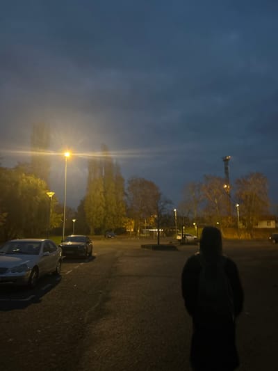 The image depicts an outdoor parking area in Haguenau, France, at dusk or night under an overcast, dark blue-grey sky. In the foreground, a single individual is seen from behind, positioned slightly right of the frame's center. This person wears dark clothing, possibly a coat or jacket, and a visible backpack, with their head obscured by a hood or dark hair. They face away from the camera.
To the left, a silver Mercedes-Benz sedan is parked in a marked space, facing left. Behind it and further down the lot, a dark-colored sedan is also parked. Multiple operational streetlights with bright yellow-orange light illuminate the scene, casting reflections and lens flares, particularly from the light source in the mid-left. Tall, slender trees, some appearing to be poplars, with mostly bare or sparsely foliated branches, are visible throughout the middle ground and background. A distinct cellular or radio tower with lights at its apex stands in the far right background, beyond the trees. The ground is asphalt, showing faint parking space markings.