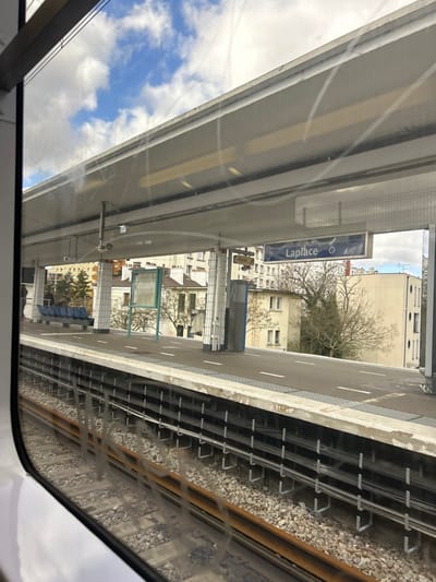 The image displays an elevated train station platform, identified as "Laplace" station in Arcueil, France, viewed from inside a train car through a window with visible smudges and scratches.
The concrete platform runs horizontally across the mid-ground. A blue sign with white lettering clearly displays "Laplace" and a train symbol. An overhead concrete canopy extends across the platform. White square-tiled columns support the canopy. On the left side of the platform, a row of blue benches is visible, with one individual partially discernible near them. A green-framed information board stands on the platform to the right of the benches.
Adjacent to the platform, two parallel train tracks are laid on a bed of gravel ballast. A metallic third rail, or similar conductor rail, runs alongside the tracks. Below the platform edge, a series of cables are secured horizontally along the concrete structure.
In the background, behind the station, multiple light-colored, multi-story residential buildings are visible. Deciduous trees, mostly bare of leaves, and some evergreen foliage are interspersed among the buildings. The sky above is partly cloudy with patches of blue. The scene is illuminated by natural light, indicating daytime.