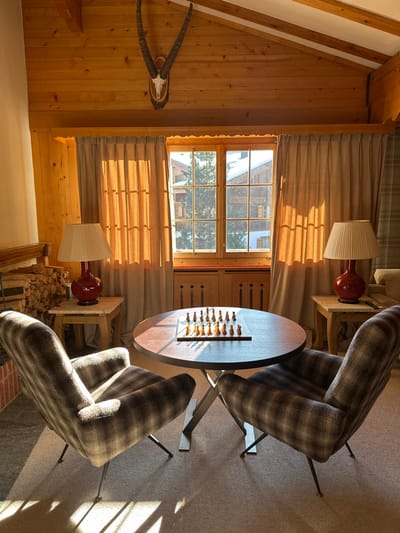 This forensic description details an interior room within an Alpine-style building, confirmed to be located in Saanen, Switzerland.
The scene depicts a well-lit living area, bathed in strong natural sunlight entering from a large multi-pane wooden window at the rear. The walls and sloped ceiling are constructed from light-toned wood paneling, featuring exposed wooden beams in the ceiling. A taxidermied animal skull with prominent curved horns, resembling an ibex or chamois, is mounted on a wooden plaque centrally on the sloped ceiling above the window.
The primary focus is a circular table with a dark wooden top and a metal X-shaped base, positioned centrally on a light beige carpeted floor. A complete wooden chess set is arranged on the tabletop, with all pieces in their starting positions. Two plaid-patterned armchairs, upholstered in shades of brown and beige, are situated opposite each other, flanking the chess table. These chairs feature slender, dark metal legs.
To the left of the window, a light-colored rustic wooden side table holds a red ceramic lamp with a pleated off-white shade. Adjacent to this, a neatly stacked pile of firewood is visible within a recessed alcove, indicating the presence of a fireplace (not fully visible). To the right of the window, an identical side table and red lamp are positioned. A section of the wall to the far right is covered with a plaid-patterned textile or wallpaper, complementing the armchairs.
The view through the window reveals snow-covered rooftops of traditional wooden chalet-style buildings and the tips of evergreen trees, consistent with a mountainous winter environment. Light beige curtains are partially drawn back on either side of the window. No individuals are present in the depicted scene.
