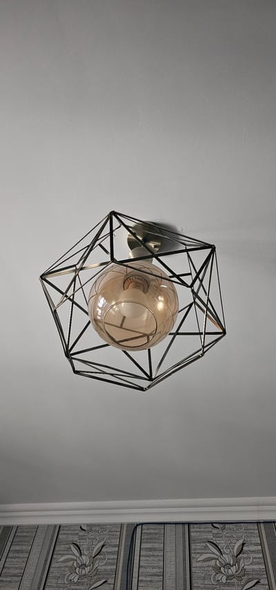 A modern, geometric ceiling light fixture hangs from a white ceiling. The fixture has a dark metal frame with angular lines, creating a cage-like structure. Inside the frame is a translucent, amber-colored glass globe that encases a single light bulb. The light fixture is centrally located in the image, with the ceiling occupying the majority of the frame. At the bottom of the image, a portion of patterned wallpaper is visible, suggesting the fixture is mounted in a room. The scene appears to be indoors, likely a residential or commercial space.
