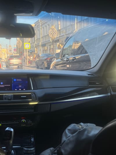 The image depicts an urban street scene in Svinoedovo, Russia, viewed from the interior of a vehicle, likely from the front passenger or driver's seat.
**Foreground (Inside Vehicle):** The dark dashboard of a modern vehicle occupies the lower half of the frame. It features a central infotainment screen displaying music information, including the track "Gold" and Russian text for "More" and "Listen on your phone." Below the screen, climate controls show a temperature setting of "20.0" degrees Celsius. A gear selector for an automatic transmission is visible on the lower left. A strip of blue ambient lighting runs across the dashboard. Part of a person's leg or lap, clad in dark, possibly leather-like material, is visible in the bottom right corner. A rearview mirror is partially visible at the top center.
**Midground (Street Scene):** The street appears to be a multi-lane thoroughfare with vehicles in multiple lanes. Directly ahead, a silver SUV, possibly a BMW X-series, with a partially visible Russian license plate (e.g., "...8 TT 777"), is positioned in the left lane. Further ahead and to its right, a dark-colored SUV coupe, resembling a BMW X6, is also visible. A black Mercedes-Benz V-Class van is parked or moving slowly to the immediate right of the viewer's vehicle, its side reflecting the surrounding buildings. A prominent yellow square sign with a black pedestrian symbol marks a crosswalk. Several pedestrians are visible on the sidewalk and near the crosswalk. Street poles are adorned with ornate, cage-like decorative light fixtures. A red and white circular traffic sign is partially visible behind the silver SUV.
**Background (Buildings & Environment):** The street is lined with multi-story, light-colored buildings (approximately 3-4 stories) featuring classical architectural details such as ornate windows and cornices. The ground floors appear to house commercial establishments. The scene is brightly illuminated by direct sunlight, casting reflections on vehicles and surfaces, suggesting a clear day.