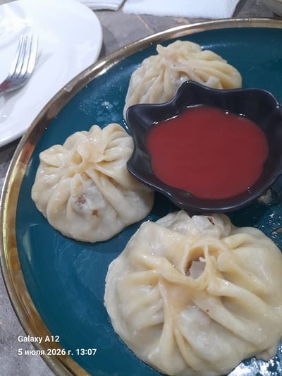 This image shows a close-up of three khinkali, a type of Georgian dumpling, served on a teal-colored plate with a gold rim. A small, black, irregular-shaped dish contains a red sauce, likely a dipping sauce for the khinkali. One khinkali is partially obscured by the sauce dish. The khinkali are pale yellow and appear to be steamed, with visible pleats forming the characteristic top knot. A fork rests on a white plate in the upper left corner of the frame. The background is blurred, suggesting an indoor setting, possibly a restaurant. The timestamp "Galaxy A12 5 июля 2026 г. 13:07" indicates the photograph was taken on July 5, 2026, at 1:07 PM. Based on information about the city of Kara-Köl City, Kyrgyzstan, this image depicts a meal likely enjoyed at a local eatery within the city.