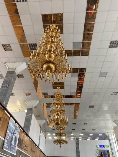 The image shows the interior of a building with a tiled ceiling featuring reflective panels. Several ornate, multi-tiered golden chandeliers are suspended from the ceiling. In the background, there are large white walls and some architectural columns clad in gray marble. A banner with text and an image is visible on the left side. On the right, a storefront sign that reads "FIX price" with a logo is present. The scene appears to be a public indoor space, possibly a shopping center or a hall. The specific location in Srednii Ikorets, Russia, is identified.
