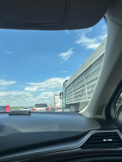 The image shows a view from inside a vehicle, looking out at a traffic jam on a sunny day. The sky is blue with scattered white clouds. In the foreground, the dashboard of the car is visible, including a vent. Ahead, there are several vehicles, including cars and trucks. One truck is particularly prominent, with a large grey cargo bed. Some of the vehicles have visible license plates or markings. The environment appears to be a highway or a major road. The scene suggests a standstill or slow-moving traffic. The location is specified as Aiuta, Russia.