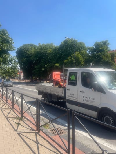 This is a photograph of a white construction van parked on a street in Stupava, Slovakia. The van is branded with "ARCHIRAUM BAU" and features construction-related services such as "STAVBA" (construction), "REKONŠTRUKCIE" (renovations), and "PROJEKTY ZÁHRAD REALIZÁCIE ZÁHRAD" (garden projects, garden implementations). The van has a flatbed on the back, carrying an orange piece of equipment.
A person is visible inside the driver's cabin. The scene is bathed in bright sunlight, suggesting it is daytime, likely a sunny day. The background shows mature green trees, a sidewalk made of paving stones, and a street with visible lane markings. A black metal fence runs along the sidewalk. The overall impression is of a business operating on a typical sunny day in a town. Text on the van includes a phone number (0915 057 708), an email address (archaumbau@gmail.com), and the company's address (Čeladice 303, 95103 Čeladice).