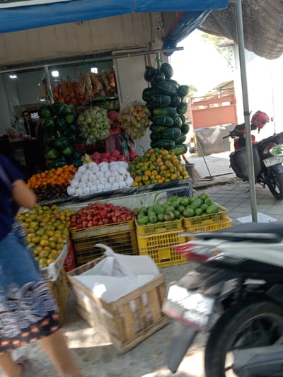 The image depicts an outdoor market stall in Kemamang, Indonesia, displaying a variety of fruits and other goods. In the foreground, a person wearing a blue shirt and patterned shorts is partially visible, walking towards the left. Behind them, a wooden crate contains white paper or plastic items, possibly packaging. To the left, several crates are filled with oranges and what appear to be limes. A large basket of red fruits, possibly tomatoes, is positioned in the center, alongside a crate of green fruits, likely avocados. Further back, a stall displays bundles of dried goods hanging from a rack, and more fruits such as watermelons and what appear to be guavas or similar small green fruits are suspended or displayed in baskets. A red helmet rests on the back of a parked motorcycle on the right side of the frame, which has a license plate with the characters "S 2952". The market appears to be under a blue tarp awning, with some sunlight filtering through.