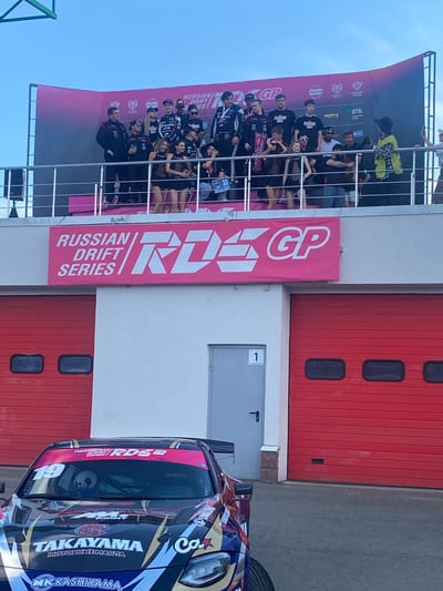 The image depicts a podium ceremony at what appears to be a motorsports event, likely the Russian Drift Series (RDS GP) in Beryozovka, Russia. Several individuals, including drivers in racing attire and women in swimwear, are standing on the podium. A large banner behind them reads "RUSSIAN DRIFT SERIES RDS GP". Below the podium, a race car is parked, adorned with various sponsor logos and the number "19". The background includes a building with red garage doors and a clear sky. The overall scene suggests a celebratory atmosphere following a competition.
