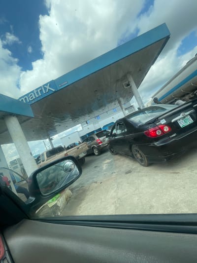 This media file captures a scene at a gas station in Ado-Ekiti, Nigeria, with multiple cars lined up, suggesting a queue for fuel. The perspective is from inside a vehicle, looking outwards.

The primary subjects are the cars, with a black Toyota Corolla prominently featured in the foreground. The license plate of this car reads "YEE 871AE". Several other vehicles are visible behind it, also waiting. The gas station structure is a prominent object, with a large, angular blue and grey canopy. The word "Matrix" is visible in large white letters on a blue section of the canopy.

The setting is an outdoor gas station with a concrete ground. The time of day appears to be daytime, indicated by the bright sky and clouds. The weather seems to be partly cloudy.

The activity is a typical gas station queue, with vehicles lined up to refuel. There are no people clearly visible within the frame of the cars or at the gas pumps, but their presence is implied by the activity.

Notable details include the visible license plate, the "Matrix" signage on the canopy, and the overall impression of a busy, functional gas station. The image is oriented vertically and rotated, which might be a result of how it was captured or processed. The presence of a side mirror from the vehicle in which the photo is taken is also a notable detail, reflecting a portion of the scene.