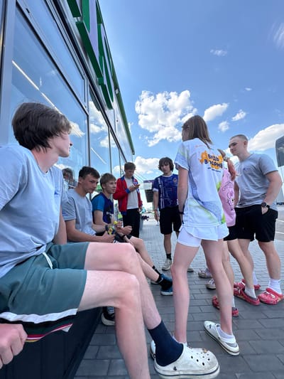 A group of at least 10 people, seemingly teenagers, are arranged in two lines, one sitting along a building facade and the other standing behind them, under a partly cloudy sky. The people in the front row are mostly wearing light-colored t-shirts and shorts. Several of them appear to be looking upwards. The people in the back row are wearing athletic attire. The building facade has large glass windows and green lettering. The ground is paved with gray bricks. The city is Казань, Russia.