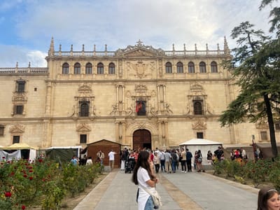 The image depicts the elaborate Plateresque facade of the Colegio Mayor de San Ildefonso, part of the University of Alcalá, situated on Plaza de San Diego in Alcalá de Henares, Spain. The multi-story building, constructed of light-colored stone, exhibits numerous ornate windows, balconies, balustrades, and intricate sculptural details including coats of arms and finials along its roofline. A Spanish flag hangs centrally from the facade.
In the foreground and middle ground, a temporary outdoor market or fair is underway, featuring various stalls erected on either side of a paved pedestrian pathway. These stalls consist of white canopy tents, dark canvas tents, and wooden structures. The pathway is bordered by decorative flowerbeds containing red rose bushes.
A moderate crowd of people, including adults and children, is present in the plaza. Individuals are observed walking, standing, and congregating around the market stalls and along the pathway. Their attire is casual and contemporary. A young woman in a white t-shirt and light-colored jeans stands in the immediate foreground, partially facing away from the camera. A young girl is partially visible in the lower right corner. A large evergreen tree partially obscures the right side of the building. The sky is overcast with visible cloud formations.