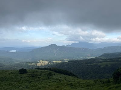 The image presents a landscape view from Illukkumbura, Sri Lanka, under a cloudy sky. The foreground is characterized by a grassy hill, dotted with small trees and bushes. The midground features a valley with dense forests and agricultural lands, with a prominent mountain in the center of the frame. Further back, a lake is visible amidst the layers of hills and mountains. The sky is covered with heavy clouds, suggesting overcast weather conditions. There are no people, subjects, or actions depicted in the view.