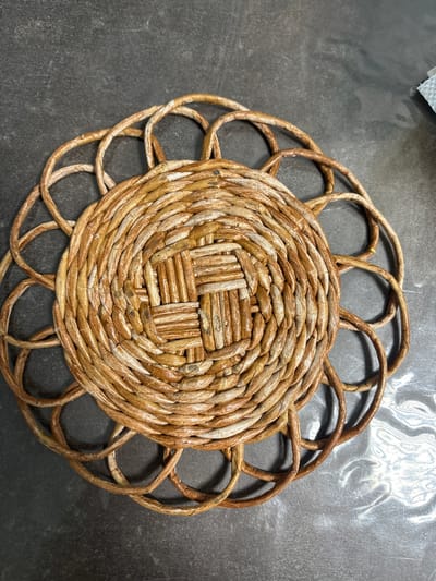 A close-up, overhead shot shows a round, woven placemat made of natural, light brown material. The placemat has a central woven square pattern, surrounded by concentric circles of woven material that extend outwards. These circles form an outer ring of decorative loops. The placemat rests on a dark, textured surface, possibly a countertop, with some light reflections and a small, white, crumpled object visible in the upper right corner. There are no people or actions depicted in the image. The location of Maidarovo, Russia is not discernible from the visual information provided.