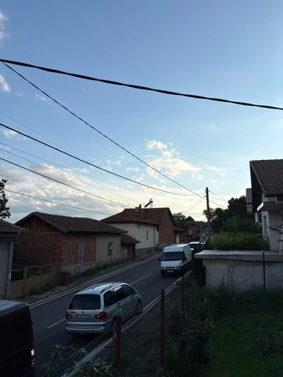 The image captures a street scene in Anton, Bulgaria, under a partly cloudy sky with a light blue hue. Multiple overhead power lines crisscross the frame, creating a sense of urban infrastructure. On the right side of the street, a modern white van is parked. Further down the road, a silver Volkswagen Sharan minivan is driving away from the camera, with the license plate "CB 0380 MX" visible. The street is lined with residential buildings, many featuring tiled roofs and brickwork. Some houses have visible solar panels on their roofs. Greenery, including trees and bushes, flanks the sides of the road and property boundaries. A metal fence runs along the right side of the street in the foreground. The overall impression is of a quiet, residential street in a small town.