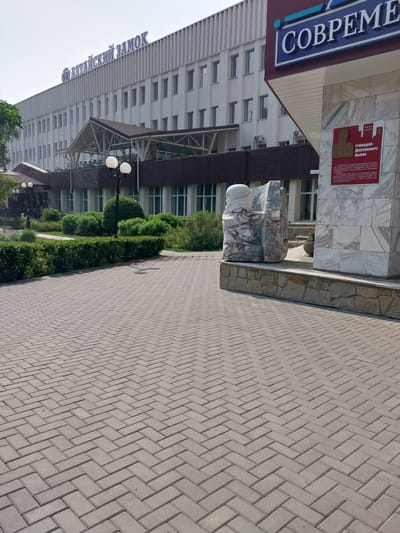 The image shows a modern building with the Russian inscription "АЛТАЙСКИЙ ЗАМОК" (Altai Castle) on its facade. To the right, a sign with the inscription "СОВРЕМЕНН" (Modern) is visible. Below this sign is a red plaque with a portrait and Russian text, indicating a historical marker related to the construction of the building. The building appears to be a hotel or resort, given its name and location. In front of the building, there is a paved walkway made of interlocking pavers, leading towards a green landscaped area with manicured bushes and trees. A sculptural element made of stone or marble is also present in the foreground. The setting appears to be outdoors on a sunny day. The image was taken in the city of Belokurikha, Russia, a known resort town in the Altai Mountains.