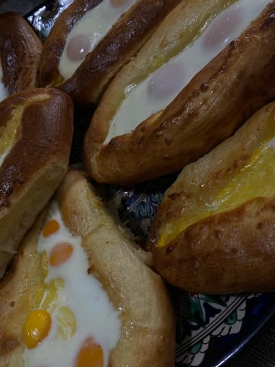 A close-up, overhead shot shows a serving of khachapuri, a traditional Georgian cheese-filled bread, on a decorative plate. The khachapuri are boat-shaped and filled with cheese and eggs, some of which are still raw, with vibrant yellow yolks visible. The bread is golden brown and appears to be slightly shiny, suggesting it may have been brushed with butter or egg wash. The plate has a dark blue base with intricate green and white floral patterns. The background is dark and slightly out of focus, with hints of a patterned surface, possibly a tablecloth. The image does not contain any people or indicate a specific location within the city of Пешнево, Russia.