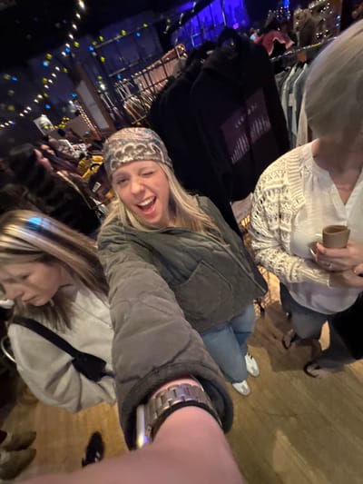 A selfie captures a group, with the foreground showing a woman wearing a patterned beanie and a quilted jacket, laughing towards the camera. Behind her, another woman in a light-colored jacket partially appears in the frame. Other people are visible in the background, some seated. The scene is lit with blue and yellow lights, creating a warm, vibrant atmosphere. A person's arm and watch are prominent in the lower right corner, indicating the angle of the shot.