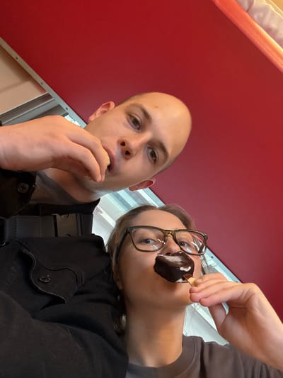 Two individuals, a male and a female, are captured in a low-angle selfie. The male, with a shaved head and visible stubble, is positioned in the upper portion of the frame, with his right hand near his mouth, appearing to eat. He is wearing a dark jacket over a grey t-shirt. The female, wearing glasses, is in the lower part of the frame, holding a chocolate-covered ice cream bar to her mouth. She is wearing a dark grey t-shirt. The background is dominated by a vibrant red surface with a white border. The lighting suggests an indoor setting, possibly a train or bus with red upholstery. The scene is devoid of any identifiable landmarks or elements that would place it specifically within the city of Bologoe, Russia.