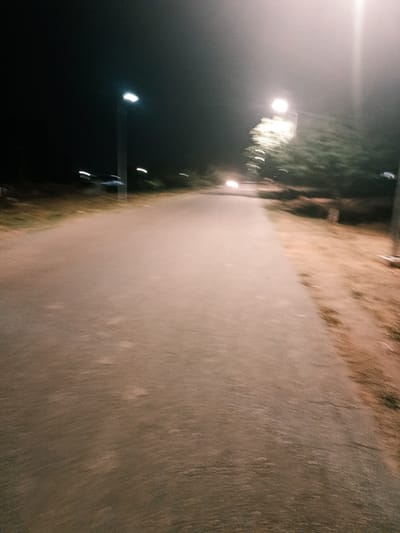 The image shows a blurry, low-light scene of a road in Rafin Pa, Nigeria. Streetlights illuminate the road, which is surrounded by vegetation and possibly trees on the sides. A vehicle with headlights is visible in the distance, moving along the road. The image appears to be taken from a moving perspective, possibly a vehicle or a person walking/running. The time is likely nighttime.