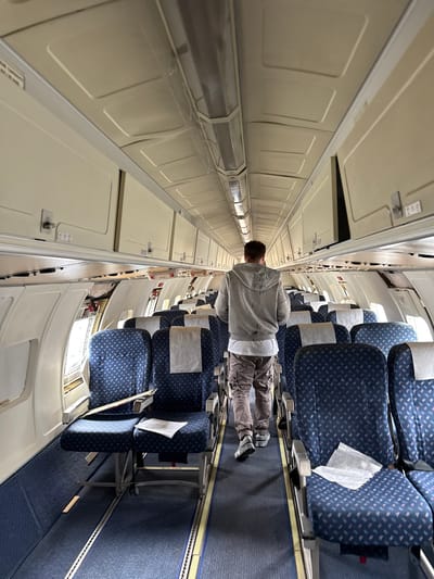 The image depicts the interior of an aircraft cabin. Rows of blue patterned seats are visible on the right side of the frame, running into the distance. A person is standing in the aisle, facing away from the camera, wearing a gray sweatshirt and khaki pants. The left side of the image shows overhead compartments with a beige or light tan color. The aircraft appears to be empty except for the person standing in the aisle. The image does not provide any information to determine the location in the city of Kraaifontein, South Africa.