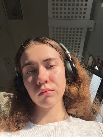 A young woman with light brown hair and headphones on is taking a selfie. She is wearing a white ribbed t-shirt. The background appears to be the interior of a vehicle, possibly a bus or train, with a grid-like pattern on the ceiling and a window on the right. Sunlight is streaming in from the right, casting shadows across her face. There is a red and white circular warning sign visible through the window. The location is identified as Винзили, Russia.