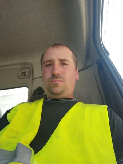 The image is a close-up, selfie-style shot of a Caucasian man inside a vehicle, possibly a truck cab. He is wearing a bright yellow safety vest over a black shirt. The man has light brown hair, is clean-shaven except for some stubble on his chin and upper lip, and has light-colored eyes. The interior of the vehicle appears to be made of grey plastic and fabric. The background shows the window of the vehicle with bright light coming through, suggesting it is daytime. There is a visible door frame on the right side of the image. The man's expression is neutral, and he is looking directly at the camera. The image provides no information about the city of Costești, Moldova.