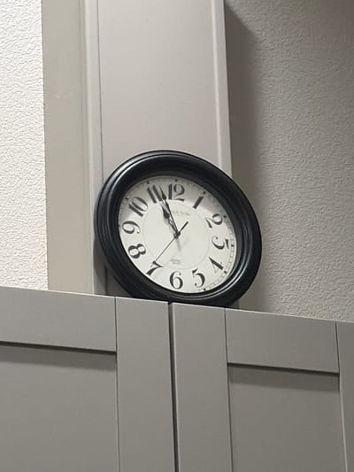 This is a close-up, eye-level shot of a black analog clock with a white face and black Roman numerals. The clock is positioned on a light-colored ledge, which is in front of a textured gray wall. To the right of the clock, a portion of a gray cabinet with horizontal paneling is visible. The clock hands are not clearly visible, making it impossible to determine the exact time. The lighting suggests it is daytime, though no specific time of day can be inferred. The environment appears to be indoors, possibly a home or office, given the presence of the clock and cabinet. There is no discernible text other than the numerals on the clock face. No people or weather conditions are visible. The image captures a simple, everyday object in a neutral setting.