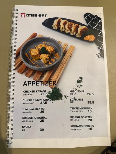 A close-up shot captures an open, spiral-bound menu from a restaurant named "ONEE-SAN," featuring an "APPETIZER" section. The menu is laid flat on a light-colored surface, likely a table. Two food items are prominently displayed as part of the menu's design: on the upper left, a black, elongated dish holds several pan-fried dumplings (gyoza) with a small dollop of sauce; on the lower center, a gray ceramic plate with a wooden trivet showcases what appears to be fried chicken pieces (possibly Chicken Karage) drizzled with a creamy sauce and garnished with fresh parsley.
The menu lists various appetizers with their names in English, along with Japanese and Indonesian translations, and corresponding prices. Visible text includes: "ONEE-SAN" as the brand name, and "APPETIZER" as the section title. The listed items are "CHICKEN KARAGE" (29.5), "CHICKEN SKIN KARAGE" (27.5), "DIMSUM MΕΝΤΑΙ" (29), "DIMSUM ORIGINAL" (25), "GYOZA" (30), "MISO SOUP" (24.5), "EDAMAME" (25.5), "TEMPE MENDOAN" (15), "PISANG GORENG" (20), and "KENTANG GORENG" (15). The currency is not explicitly stated but is likely Indonesian Rupiah given the location context of Kecamatan Cisaat, Indonesia. The menu design is clean, with the items and prices clearly legible, indicating a modern dining establishment.