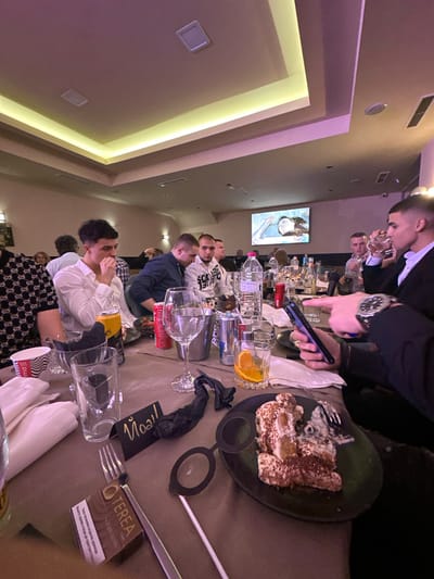 The image shows a gathering of people at a table, possibly in Stoikite, Bulgaria. The setting appears to be a restaurant or event space with modern lighting fixtures on the ceiling. A group of people are seated around a long table, interacting with each other. A man is using a mobile phone, while food and drinks are present on the table, including a plate with desserts, glasses of water and other beverages, bottles of various liquids, and a pack of tobacco product, TEREA. Several glasses and napkins are also arranged on the table. The atmosphere seems convivial, suggestive of a social gathering. A painting can be seen on the wall.