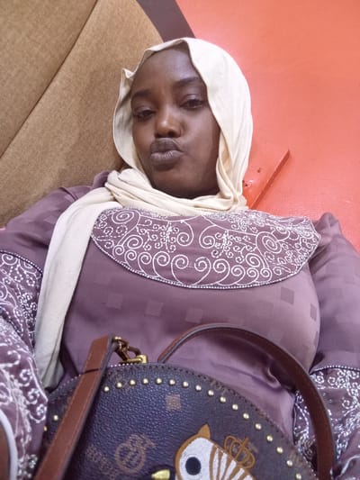 A medium shot depicts a woman wearing a light beige headscarf, a mauve dress with white patterned trim, and a dark-colored, studded purse. The woman is seated, leaning against a tan-colored surface with an orange wall visible in the background. Her lips are pursed. The location is in the city of Ajiwa, Nigeria.