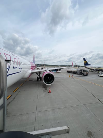 The image shows an outdoor scene at an airport, likely Balice, Poland, on a cloudy day. The foreground features the side of a white airplane with pink and blue markings, including the Wizz Air logo on its fuselage and a pink engine cowling. The airplane is parked on a tarmac, with yellow and black painted lines visible on the ground.
In the mid-ground, other aircraft are parked. To the right, a Ryanair airplane with a blue and yellow tail is visible. Between the two aircraft, a fuel truck with "LOTOS Aviation" branding is parked, suggesting refueling operations are underway. Other vehicles, including a bus, are also present on the tarmac.
In the background, more aircraft and airport infrastructure, such as lighting masts, are visible, along with a line of trees and hills in the distance. The sky is overcast with scattered clouds. The overall impression is of an active airport environment.