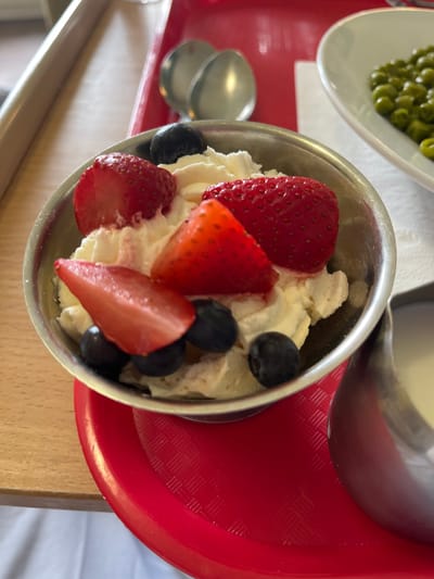 A close-up shot shows a metal bowl filled with whipped cream, strawberries, and blueberries. The bowl is placed on a red tray. In the background, a white napkin and a bowl of green peas are visible. The scene appears to be indoors, possibly in a cafeteria or restaurant. The image does not provide any information about the city of Tyrrellspass, Ireland.