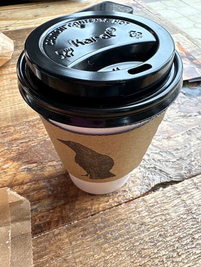 This is a close-up, overhead, rotated shot of a black coffee cup with a cardboard sleeve featuring a black owl silhouette logo. The cup is resting on a textured wooden surface. The lid of the cup is black and has embossed text that reads "CAUTION CONTENTS HOT" in a circular pattern around the rim. There are also recycling symbols visible on the lid. The cardboard sleeve is a light brown color and is wrapped around the body of the cup. The owl logo is a simple, stylized silhouette.
The scene appears to be casual, possibly a cafe or an outdoor setting, given the wooden surface which could be a table or a counter. There are no people visible in the frame, and the focus is entirely on the coffee cup and its immediate surroundings. The lighting suggests it could be daytime, but without more context, the time of day is not precisely determinable. The image is sharp, with good detail on the texture of the wood and the embossed text on the cup lid. The overall impression is one of a simple, everyday object.