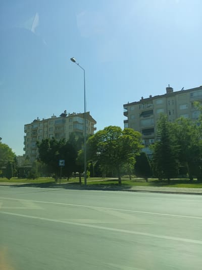 The image shows a view from a car window on a sunny day, likely in the afternoon. The scene is a city street in Karatay, Turkey, with apartment buildings and trees lining the road. In the foreground, a wide, paved road with white lane markings is visible, blurred due to the motion of the car. Beyond the road, a grassy area with trees and a few streetlights is present. Two multi-story apartment buildings with balconies and visible solar panels on their roofs dominate the background. The sky is a clear, bright blue, indicating fair weather. No people are clearly visible, and no specific event appears to be taking place. The overall impression is of a calm, everyday urban scene in a residential area.