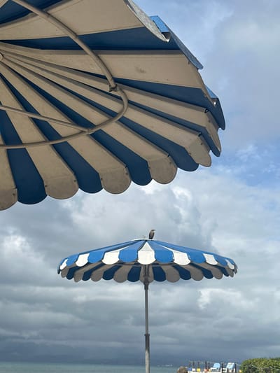 Two blue and white striped umbrellas with rounded edges stand against a cloudy sky and a distant shoreline in Le Gosier, France. The umbrella on the left is closer to the viewer, showcasing its metal frame and detailing. The second umbrella is further away, revealing its pole and the beach area.