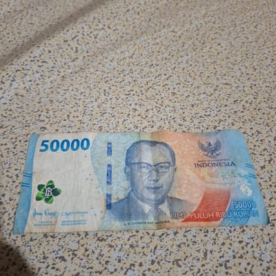 The image shows a 50,000 Indonesian Rupiah banknote lying on a textured, speckled surface, likely a table or countertop. The banknote features a portrait of Ir. H. Djuanda Kartawidjaja, the Garuda Pancasila emblem, and the denomination "50000" printed in large blue numerals. Text on the banknote includes "NEGARA KESATUAN REPUBLIK INDONESIA" and "LIMA PULUH RIBU RUPIAH." Signatures of the Governor of Bank Indonesia and the Minister of Finance of the Republic of Indonesia are also visible. There is no information in the image to determine its location in the city of Reudeuep, Indonesia.